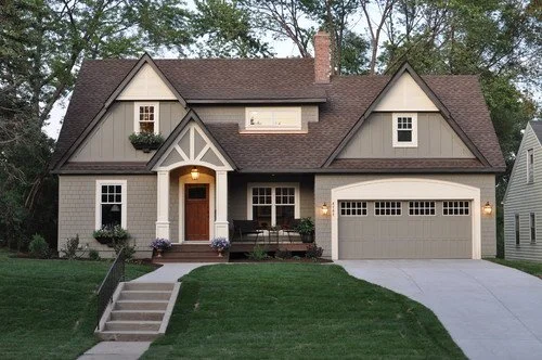 A two-story suburban house with a brown roof, beige and gray exterior, a front porch with steps, a garage, and surrounded by trees.