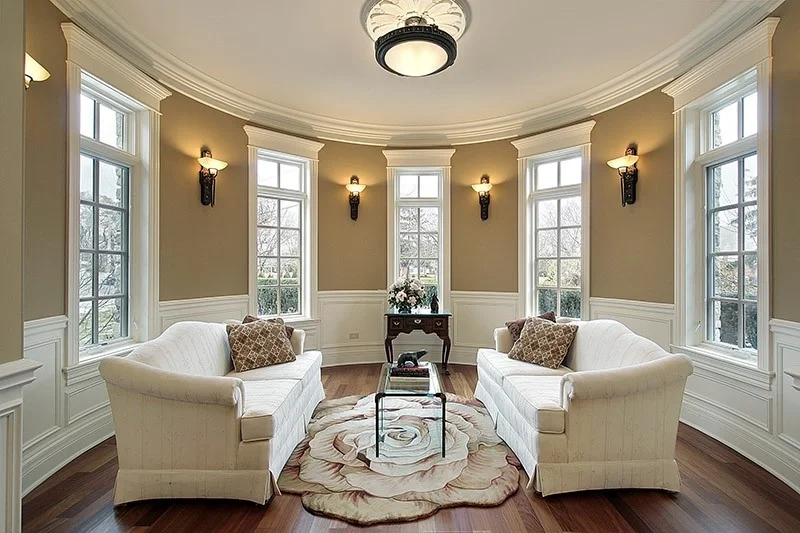A bright living room with multiple large windows, two white sofas with decorative pillows, a small table with a flower arrangement, a patterned rug, and a ceiling light fixture.