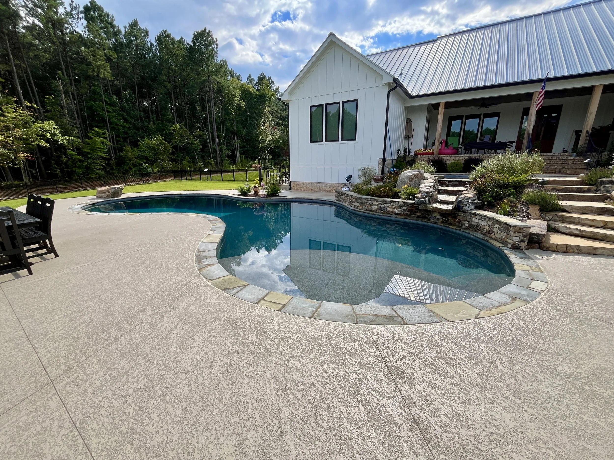 Backyard with a kidney-shaped swimming pool, concrete deck, a white house with a metal roof, landscaped steps, and surrounding trees.