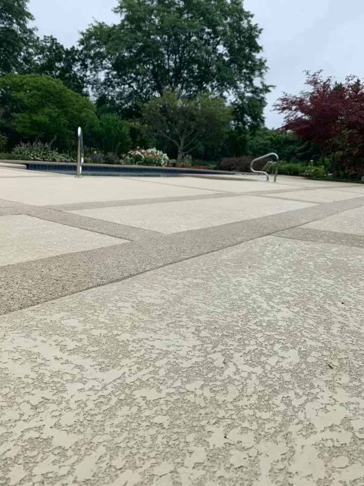 Empty poolside area with textured concrete and metal handrails, surrounded by trees and shrubs in a garden setting.