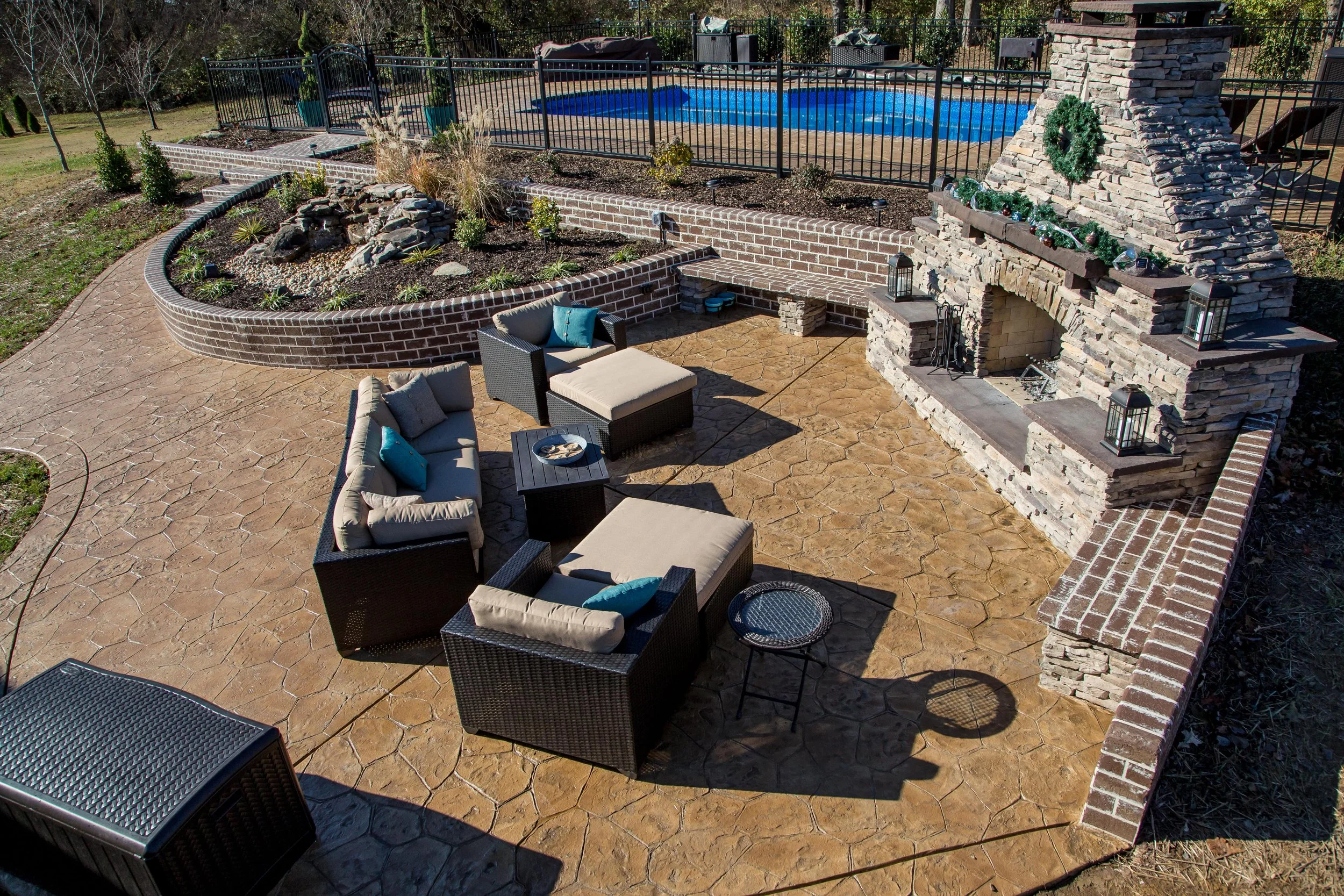 An outdoor patio area with beige furniture, a stone fireplace decorated with a green wreath and Christmas ornaments, a raised brick garden bed filled with plants, a swimming pool surrounded by a black fence, and a stamped concrete surface.