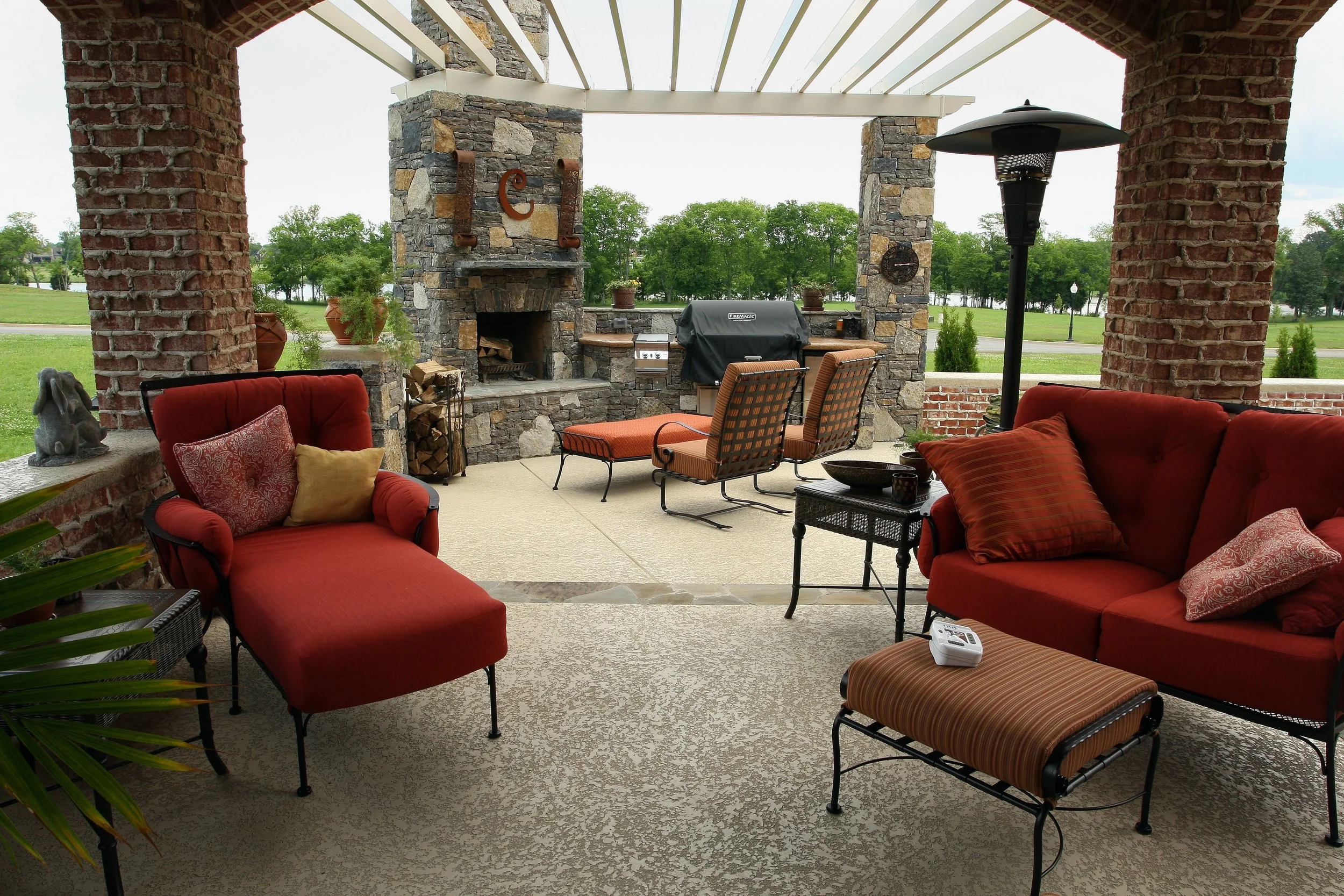 Covered outdoor patio with red and orange seating, a stone fireplace, and a view of a grassy area with trees.