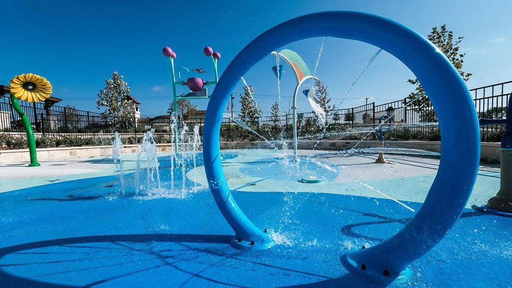 In a playground water park, there's a large blue circular splash feature with water shooting out from the edges, and colorful flower and animal-shaped water sprayers, all under a clear blue sky.