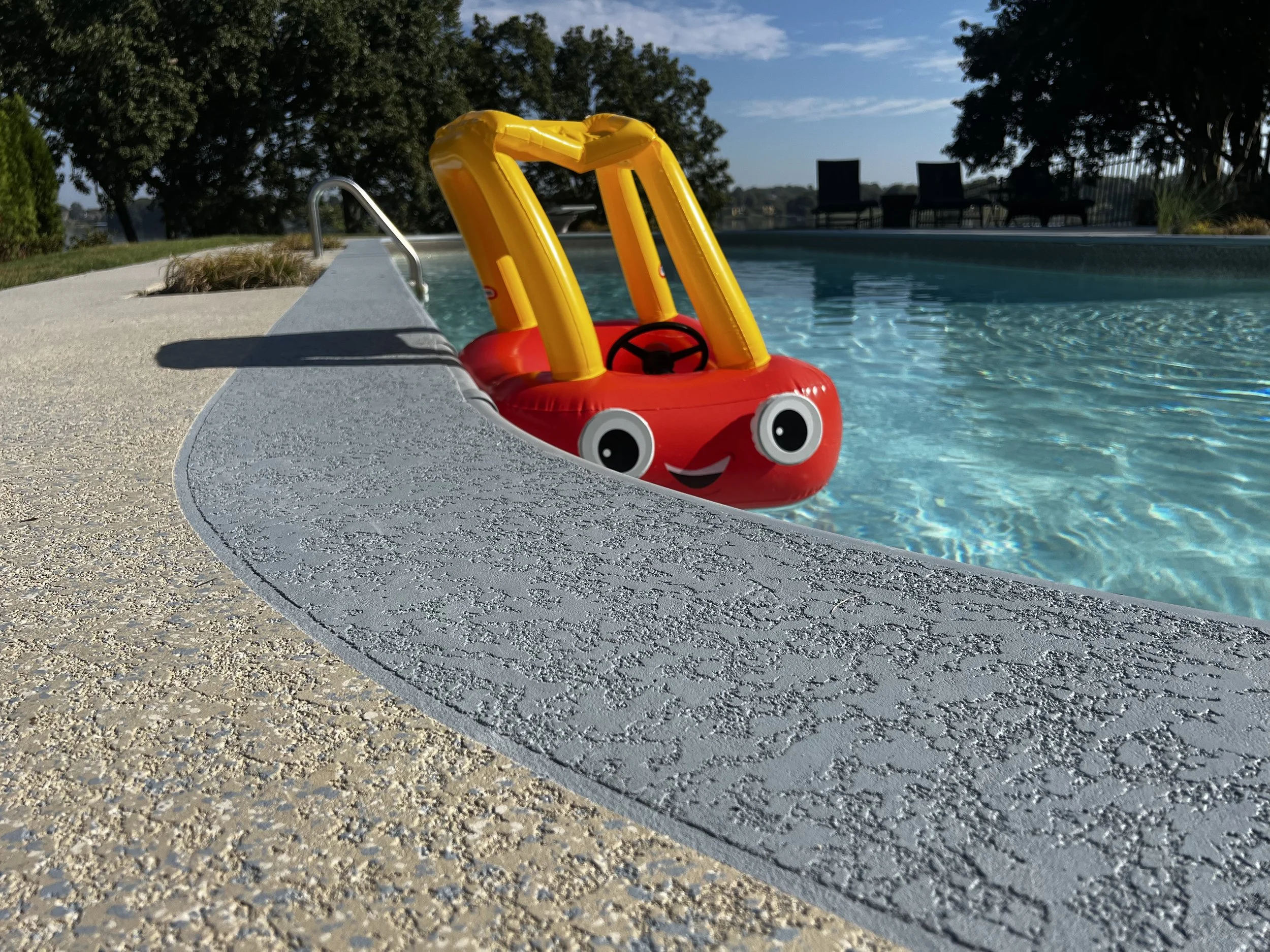 A red and yellow inflatable boat with a smiling face is floating in a swimming pool. The pool has a light-colored textured concrete deck, with trees and outdoor chairs in the background under a blue sky.