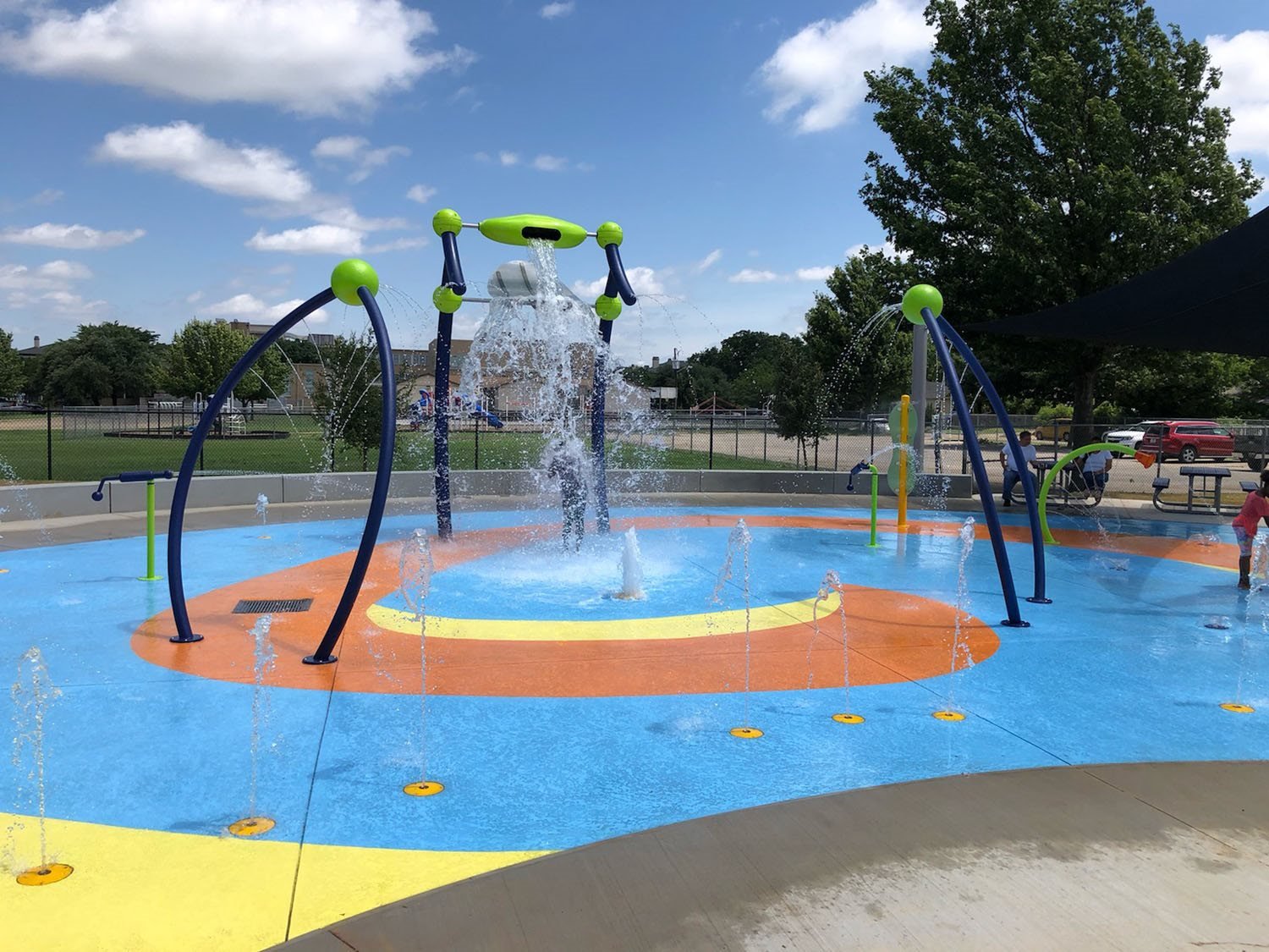 Colorful splash pad with water sprays, fountains, and spray nozzles in a park on a sunny day, surrounded by children playing and trees in the background.