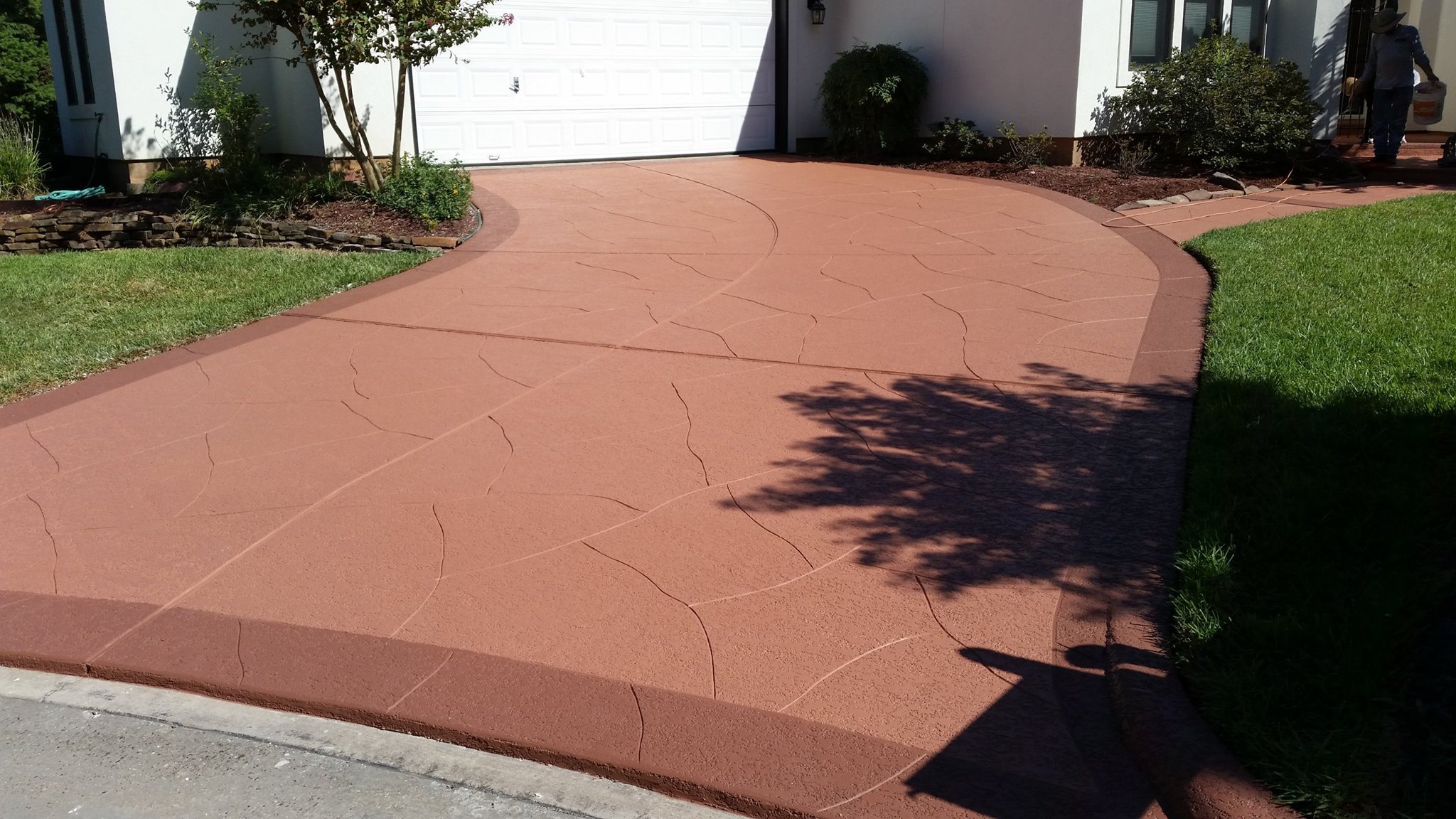 Residential front yard with a newly paved red concrete driveway, bordered by a grassy lawn and landscaped flower beds, with a person standing near the house's front door.