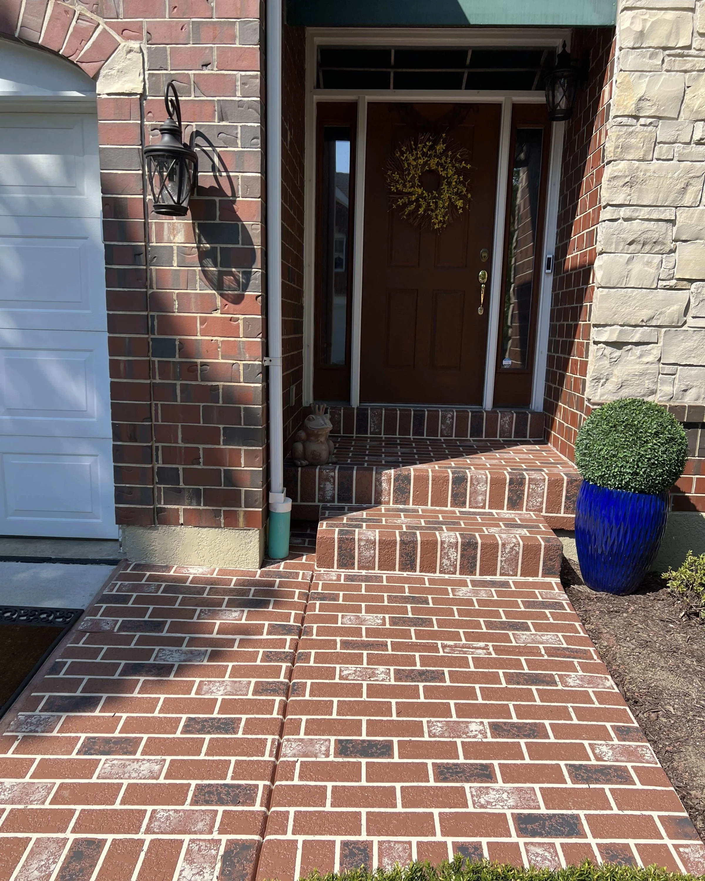 Front porch with brick steps leading to a brown wooden door decorated with a wreath. The porch has two black lantern-style wall lights, a small owl statue, and a blue pot with a trimmed green shrub. The house has brick and stone exterior walls, and a white garage door is partially visible on the left.