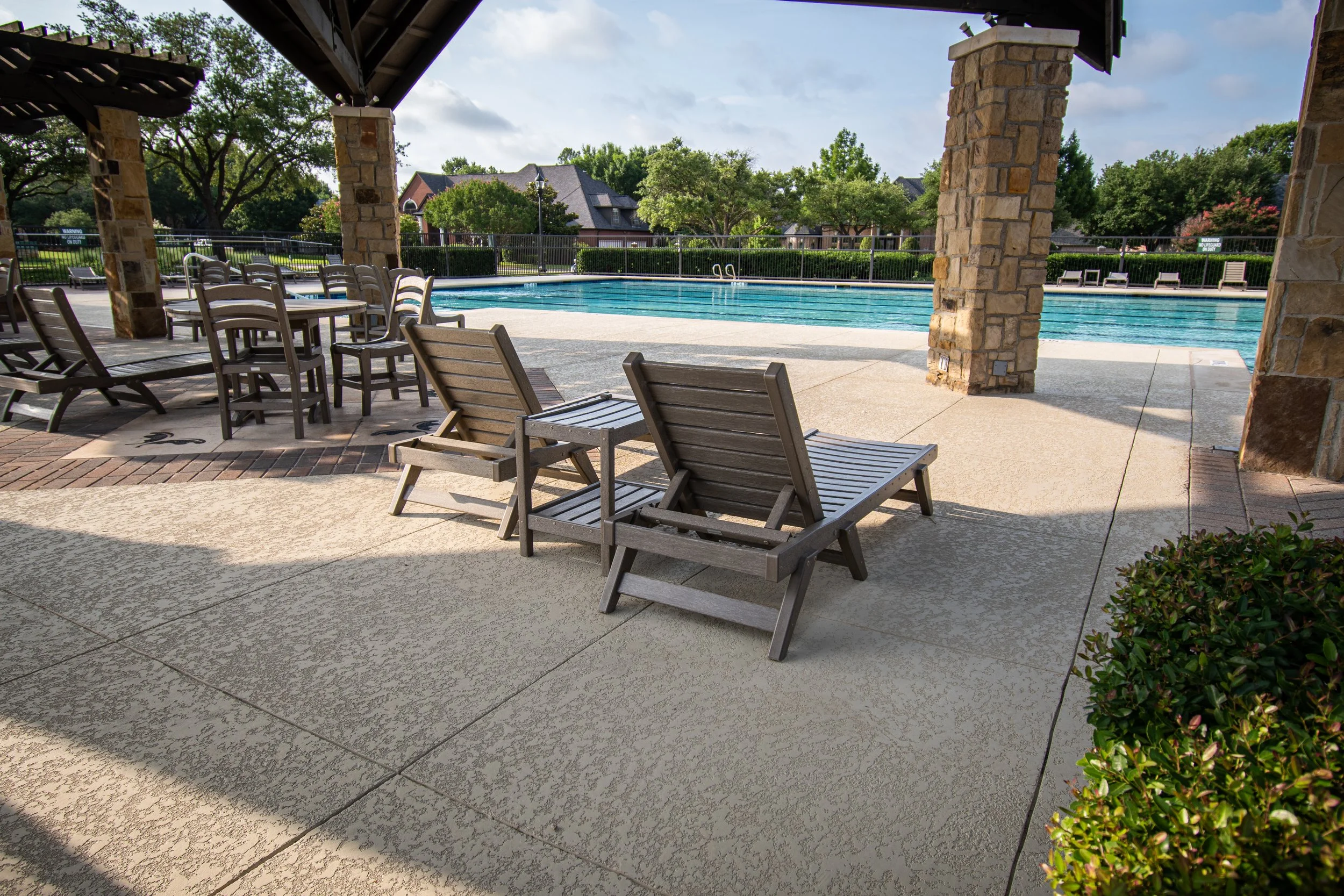 Empty swimming pool with lounge chairs and tables under a shaded patio, surrounded by trees and residential houses in the background.