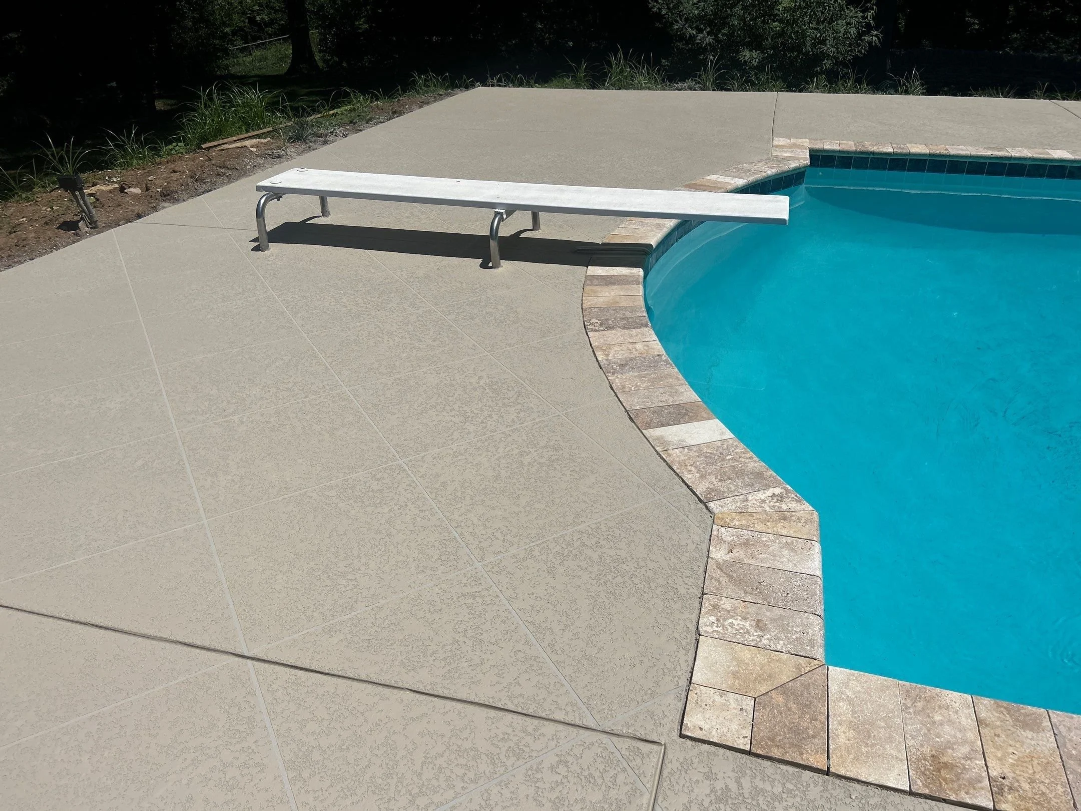 An empty swimming pool with a ladder at the corner, surrounded by beige tiles and a brick border, with a grassy area and trees in the background.