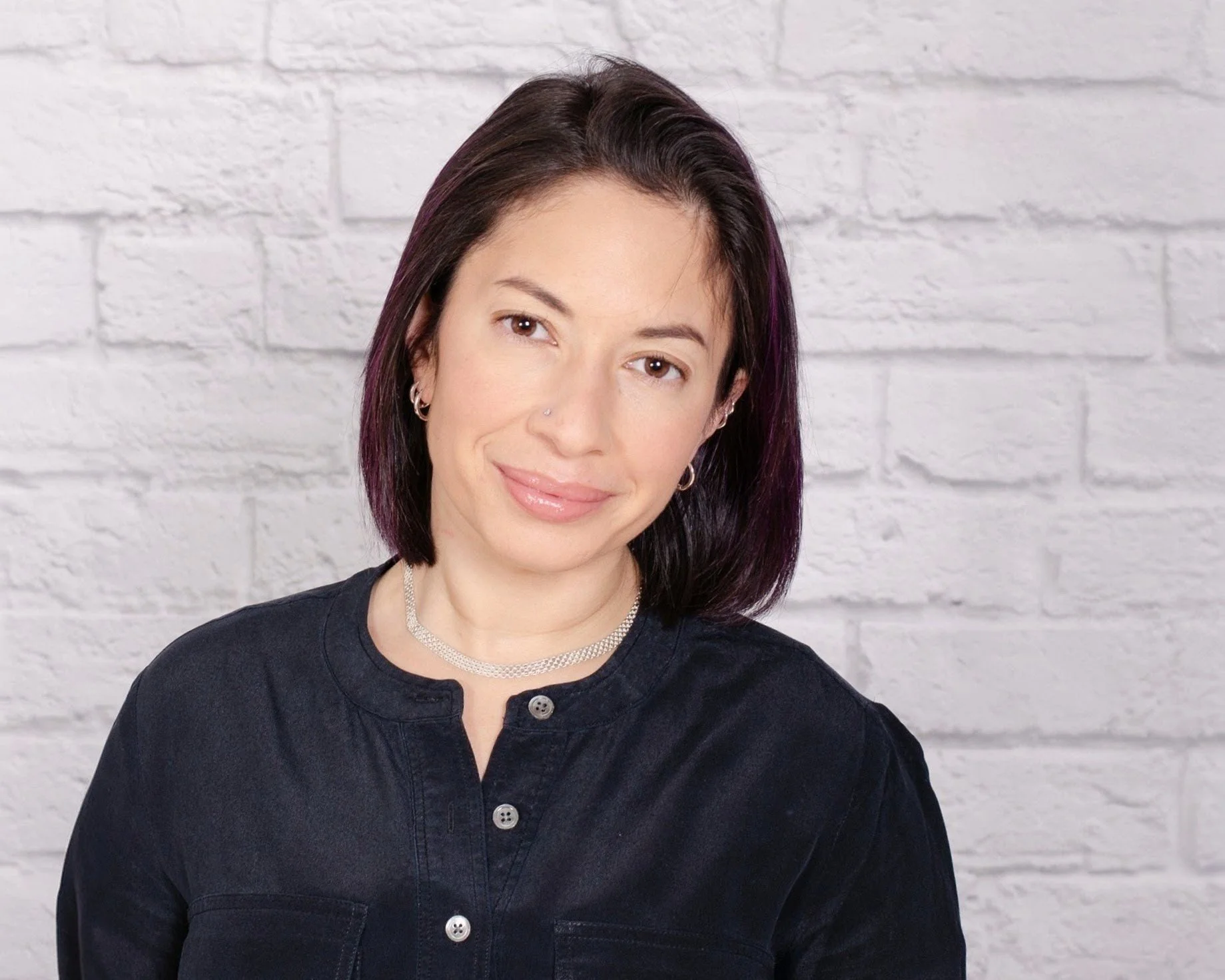 A woman with shoulder-length dark hair with purple streaks, wearing hoop earrings, a nose ring, a choker necklace, and a black shirt, smiling in front of a white brick wall.