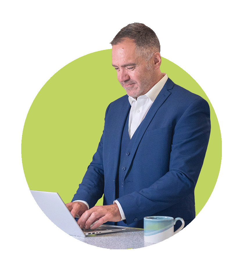 A man in a blue suit and white shirt working on a laptop at a desk with a coffee mug, standing in front of a green background.