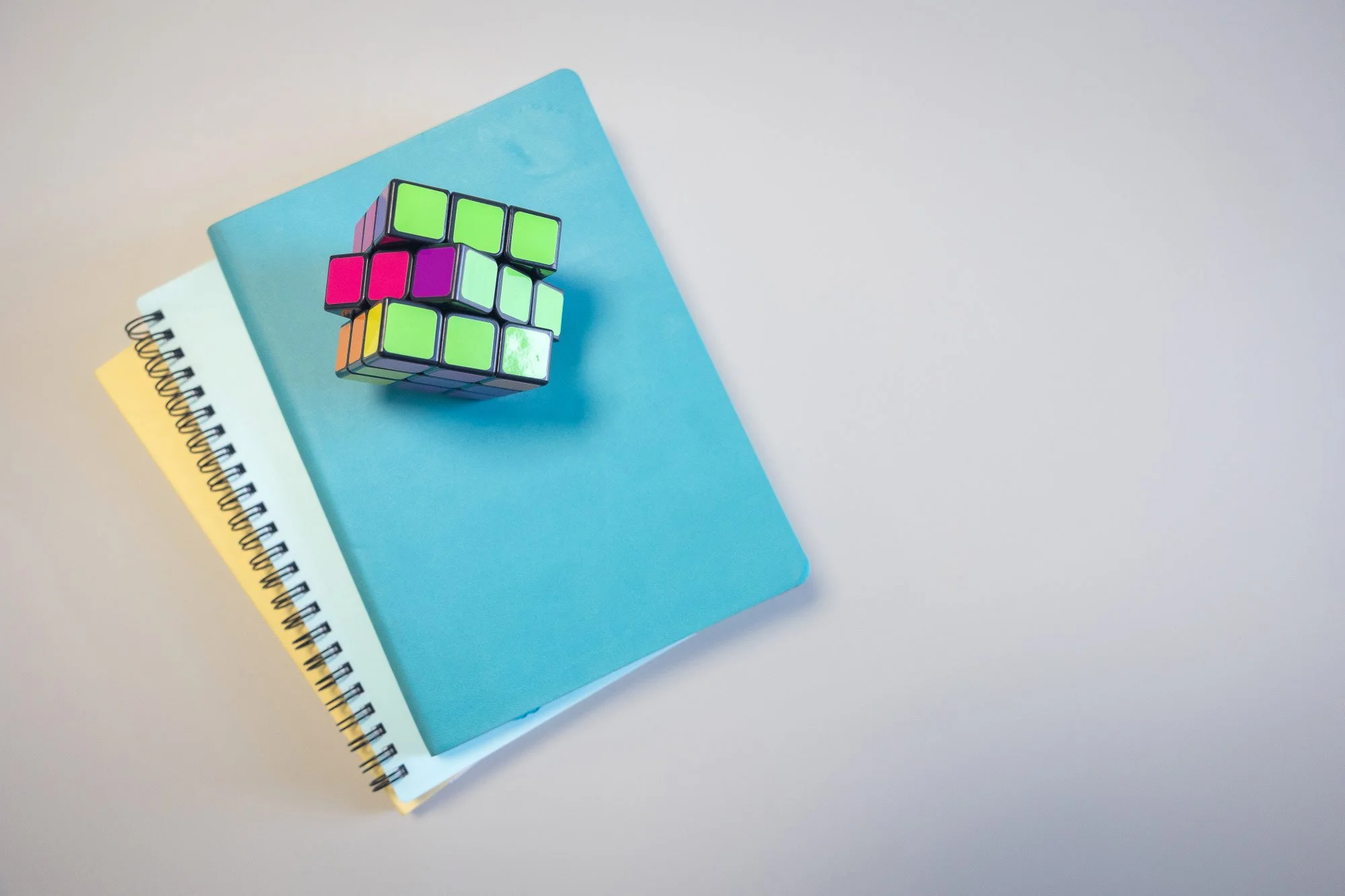 A partially solved Rubik's cube with neon green, pink, purple, orange, yellow, and white sides on top of three stacked notebooks in yellow, white, and blue.