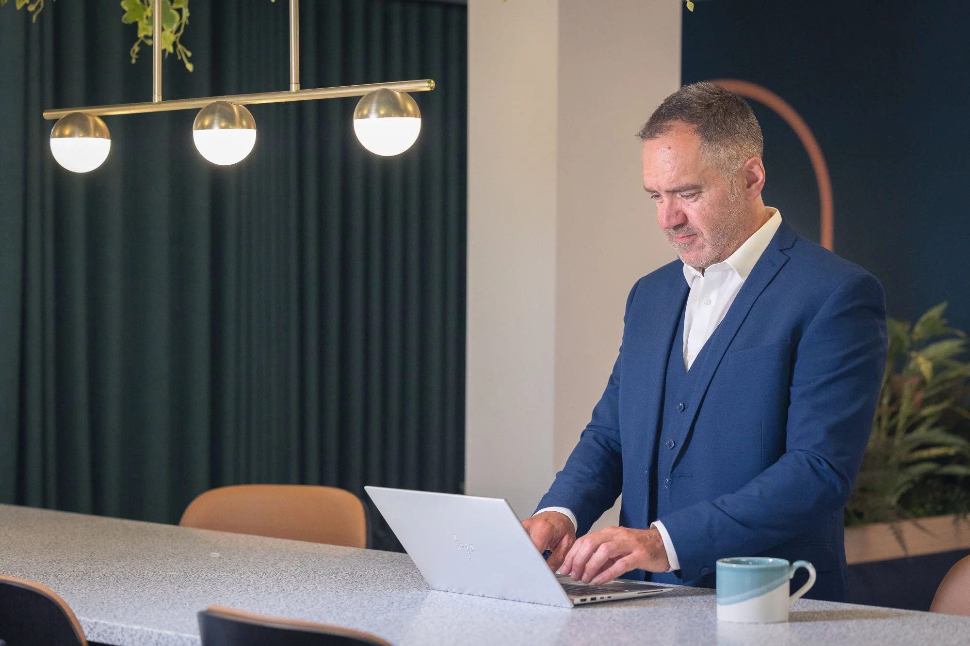 A man in a blue suit and white shirt working on a laptop at a meeting table with a green curtain in the background and a coffee mug on the table.