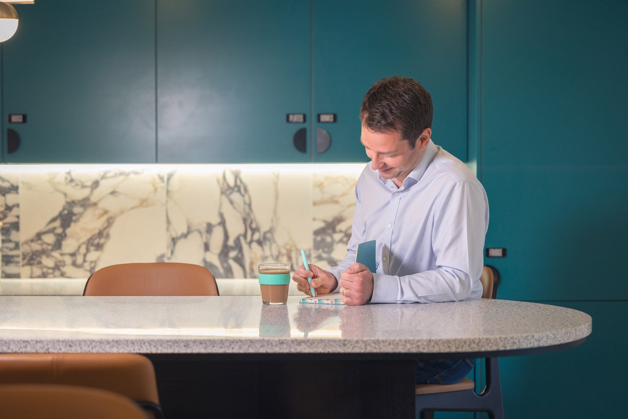 A man in a white shirt is sitting at a counter, writing on a notebook or planner with a blue pen. There is a coffee cup with a teal band on the counter. The background features a marble backsplash and teal-colored cabinets in a modern kitchen or cafe setting.