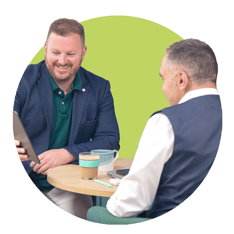 Two men sitting at a small round table having a conversation, with coffee cups and a tablet on the table, against a lime green background.