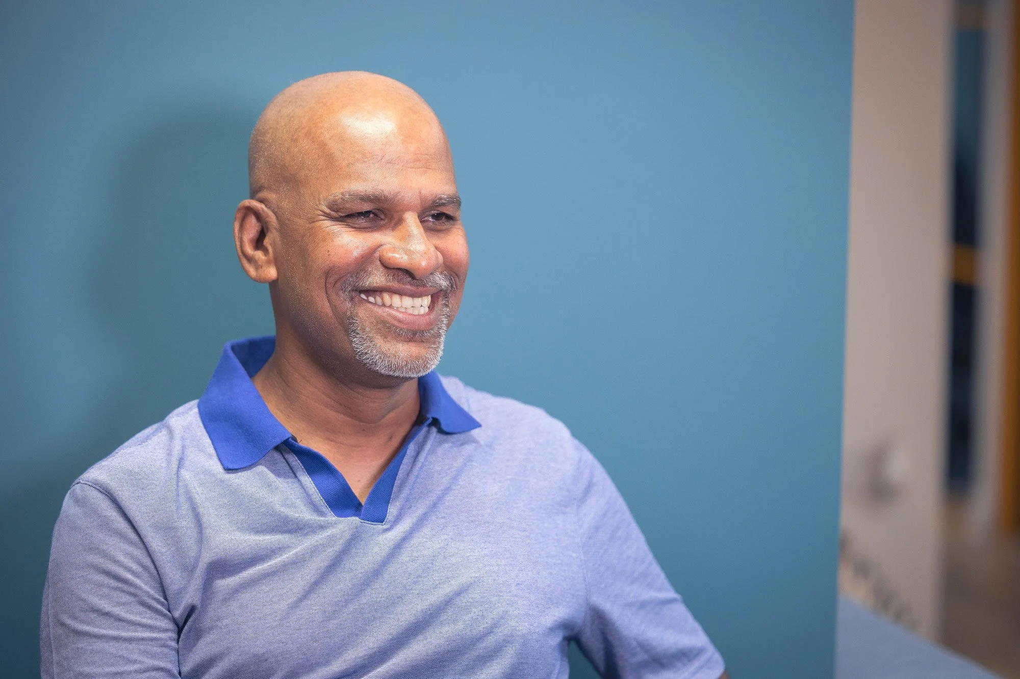A middle-aged man with a shaved head and beard smiling, wearing a blue and grey collared polo shirt, standing against a blue wall.