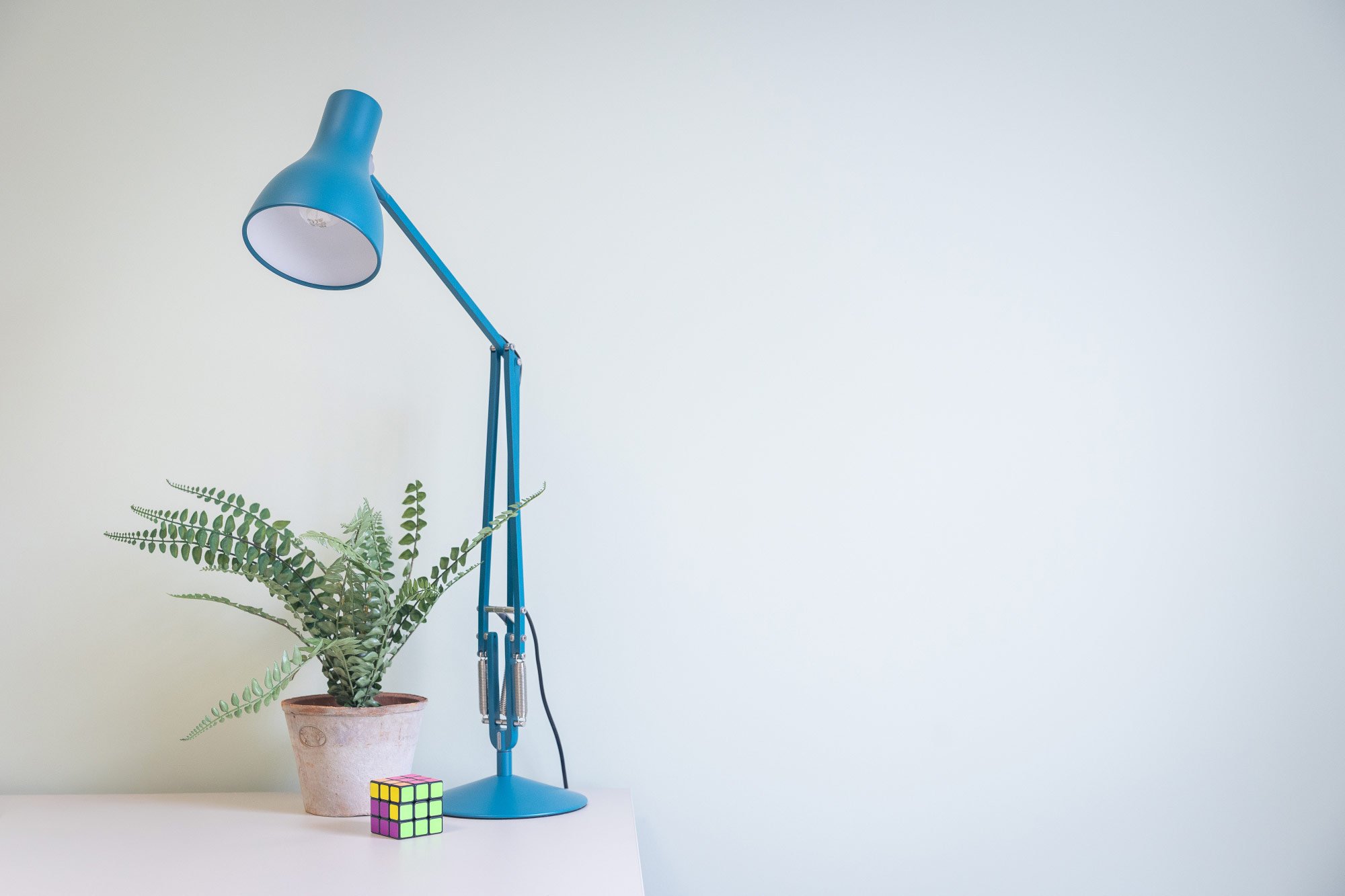 A blue desk lamp, a potted fern, a Rubik's cube, and a white table against a plain background.