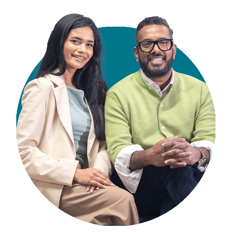 A smiling woman with long black hair sitting next to a man with glasses and a beard, both dressed casually against a blue background.