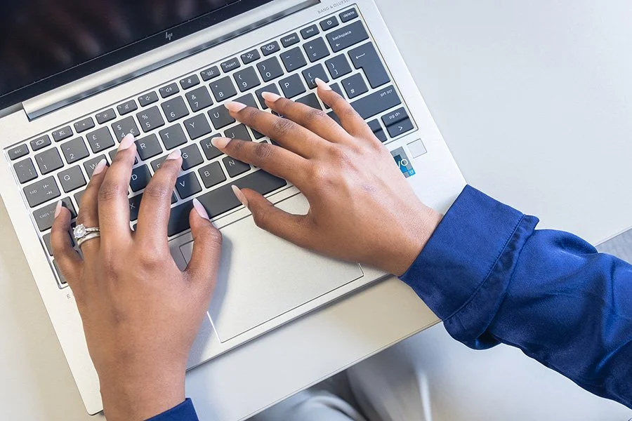 A person typing on a laptop keyboard, wearing a dark blue long-sleeve shirt, with a ring on their left hand.