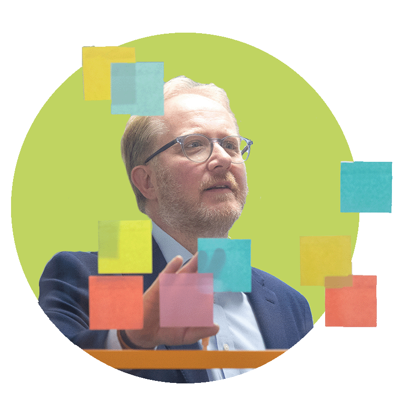 A man with glasses, a beard, and blonde hair in a suit, writing on colourful sticky notes on a transparent board against a green background.