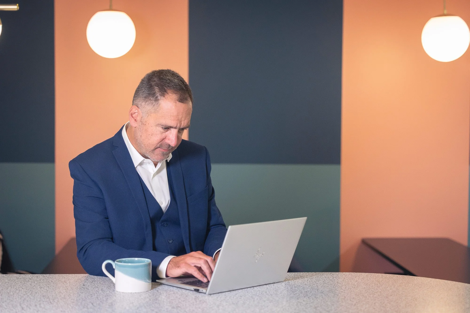 A man in a blue suit working on a laptop at a table with a mug and a cup in front of him, in a room with wall-mounted lights and multicolored wall panels.