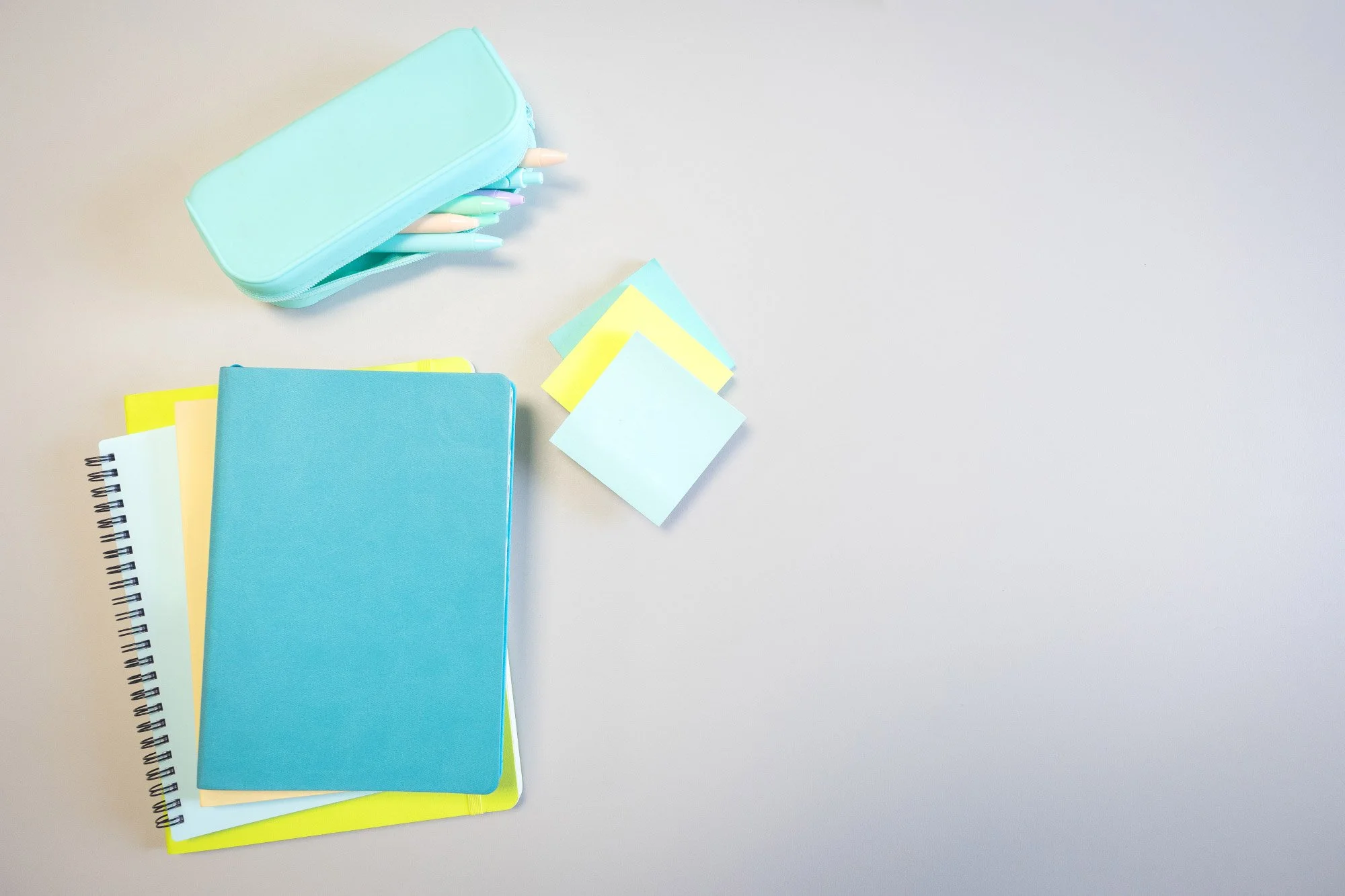A light blue pencil case containing pastel-coloured pens and pencils. Next to it are a closed blue notebook, a yellow folder, a white spiral notebook, and some sticky notes in blue, yellow, and green.