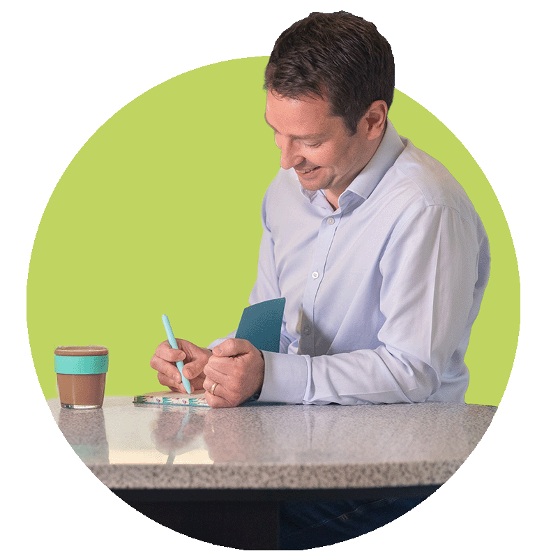 A man wearing a light blue shirt sitting at a table, smiling while writing in a notebook with a pen. There is a coffee cup on the table and a green background.