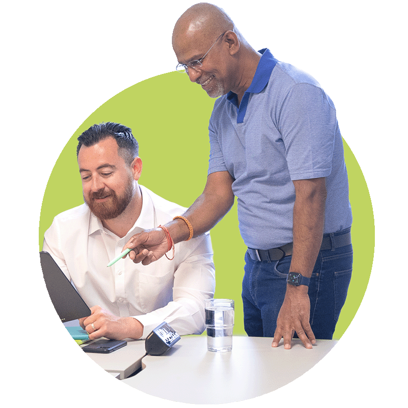 Two men in an office setting, one seated and the other standing, looking at a digital tablet. The standing man is pointing at the tablet, with a glass of water and a telephone on the table. The background is a solid green circle.