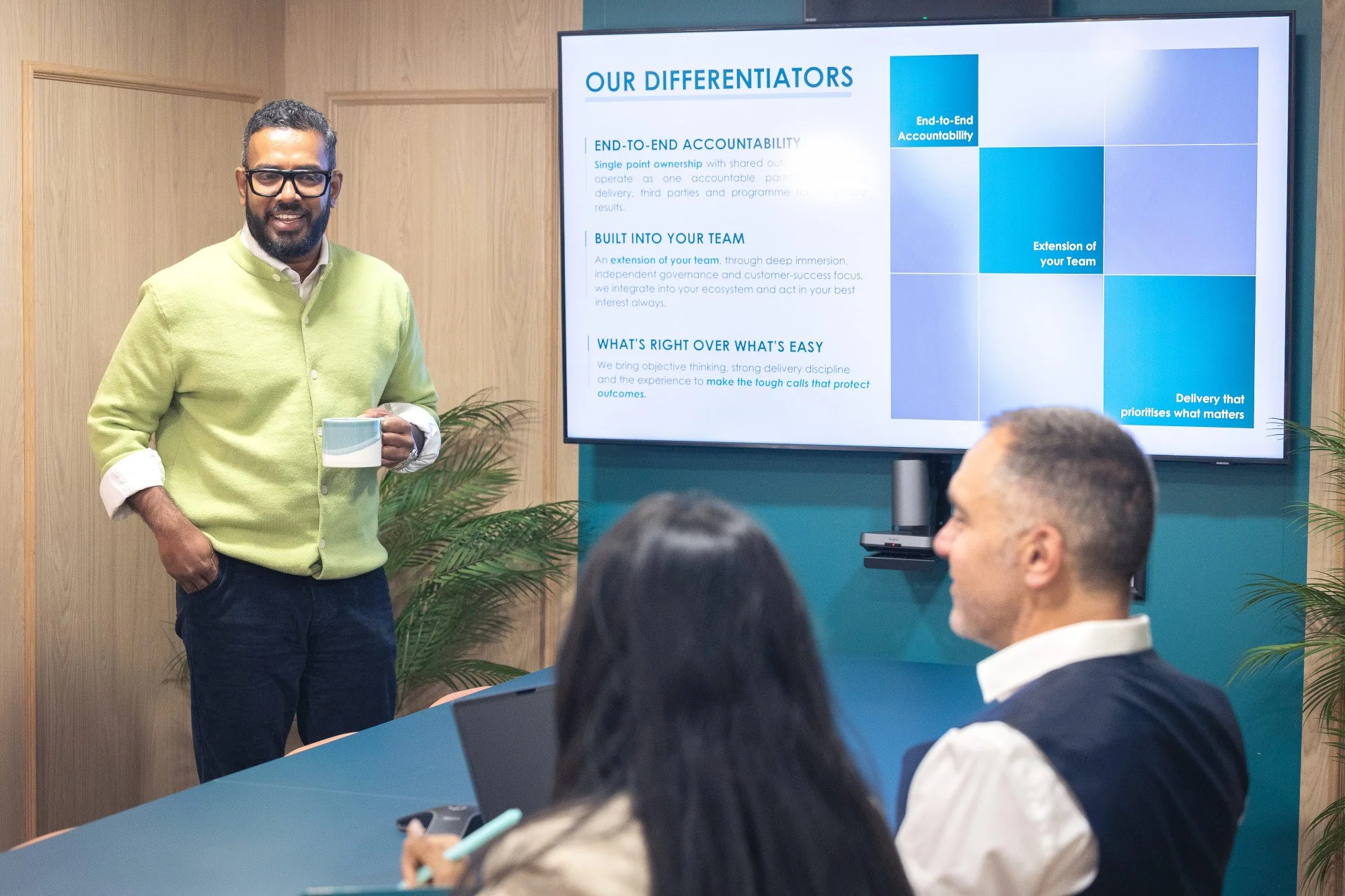Man with glasses and a beard, wearing a lime green sweater, standing in front of a presentation screen, holding a mug. Several people are seated at a table, with one person in the foreground taking notes or using a device. The presentation slide on the screen discusses team accountability and delivery.