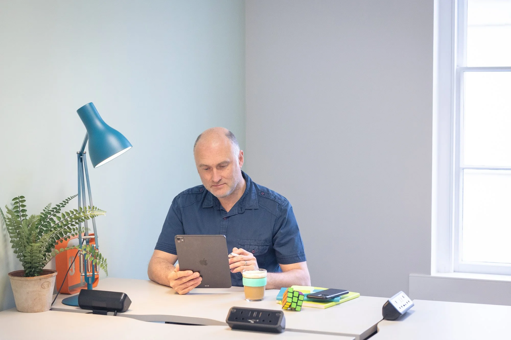 A man sitting at a white desk using a tablet. The desk has a potted fern plant, Rubik's Cubes, a smartphone, and a glass of coffee. There is a blue desk lamp, and a window with white blinds in the background.