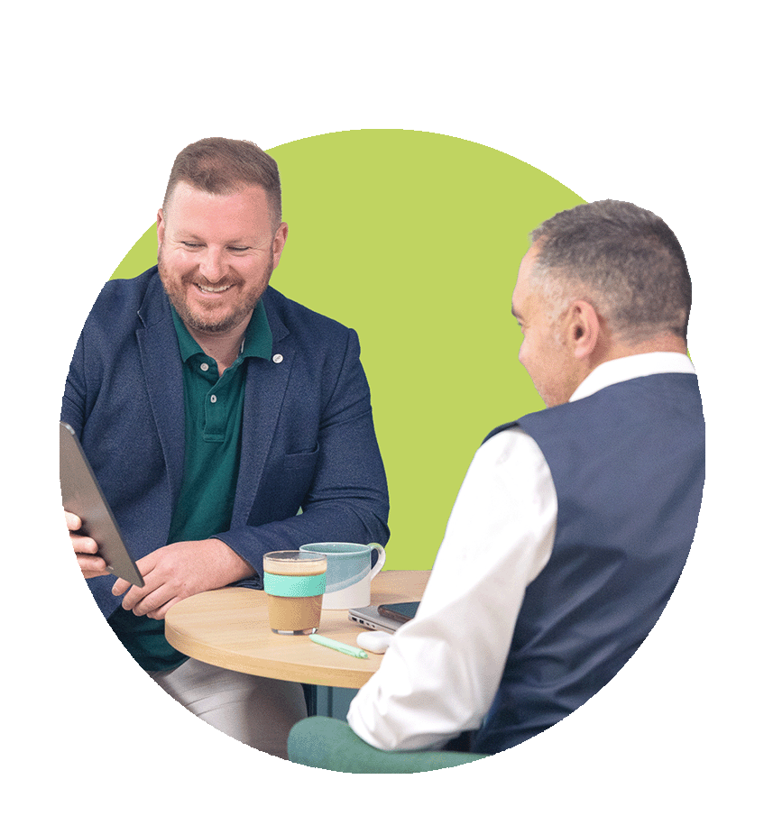 Two men sitting at a round table, smiling and talking. One is showing something on a tablet to the other. The background has a green semi-circular shape.