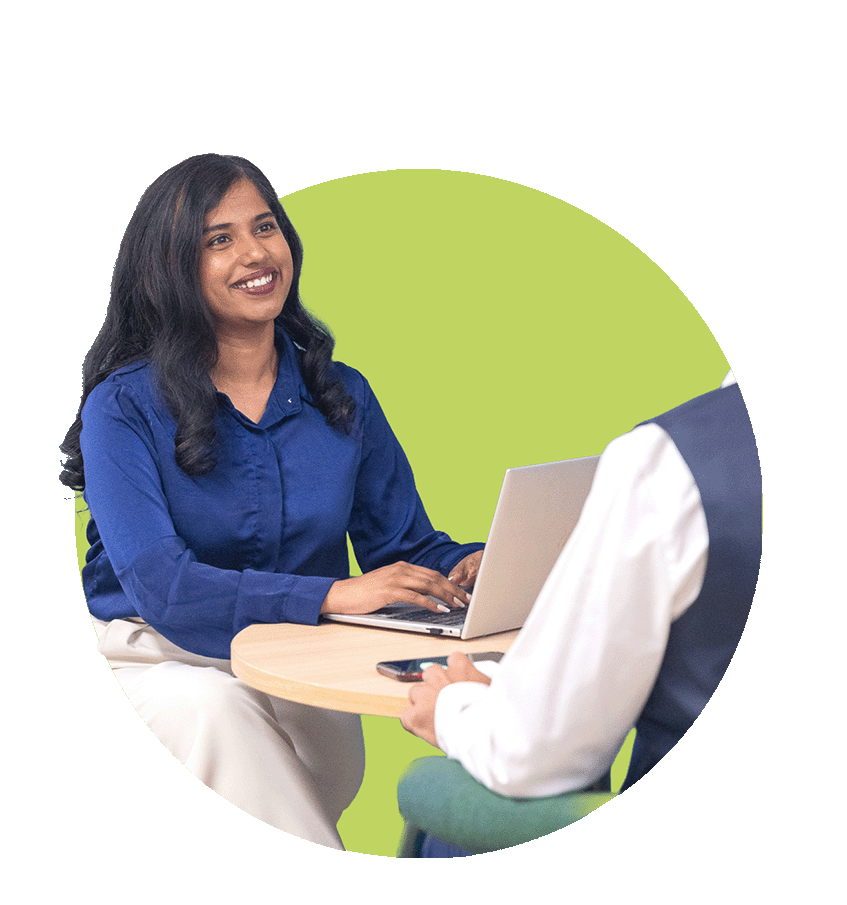 Two women sitting at a round table having a conversation, one with a laptop; woman facing the camera smiling, wearing a blue shirt, dark hair, while the other face is not visible, holding a phone, with a green background.