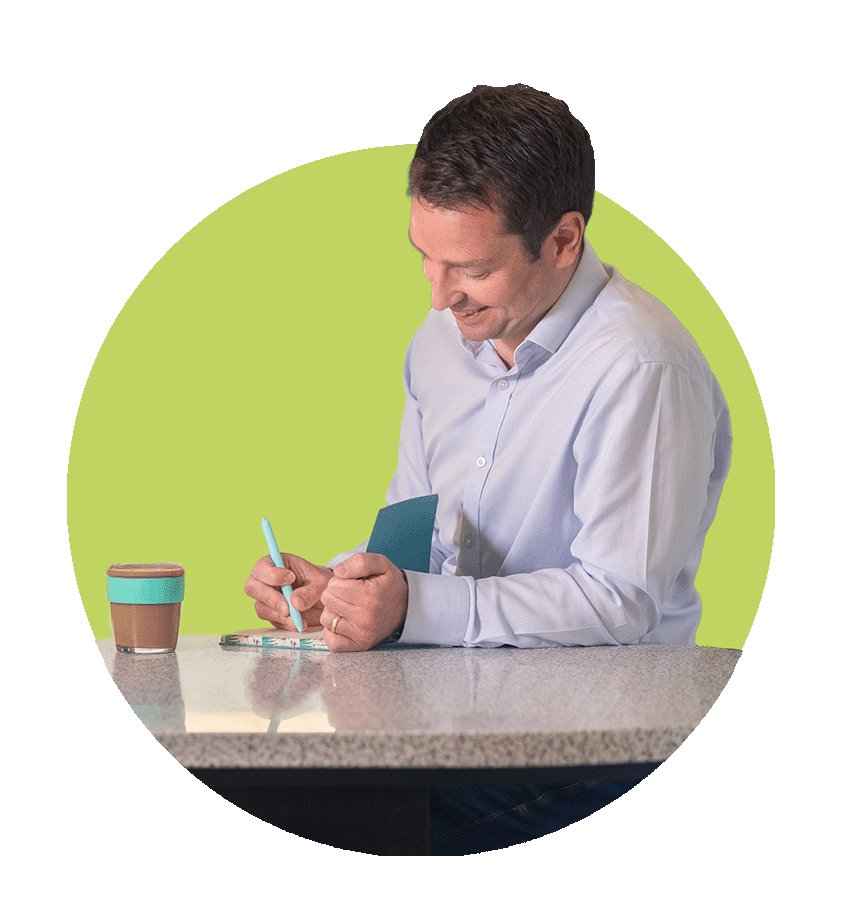 Man in light blue dress shirt sitting at a table, smiling, writing in a notebook, with a coffee cup nearby, in front of a green background.