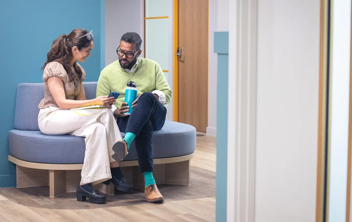 Two people sitting on a curved couch in an office, looking at a smartphone together. One is a woman with long hair, the other is a man with glasses, beard, green sweater.