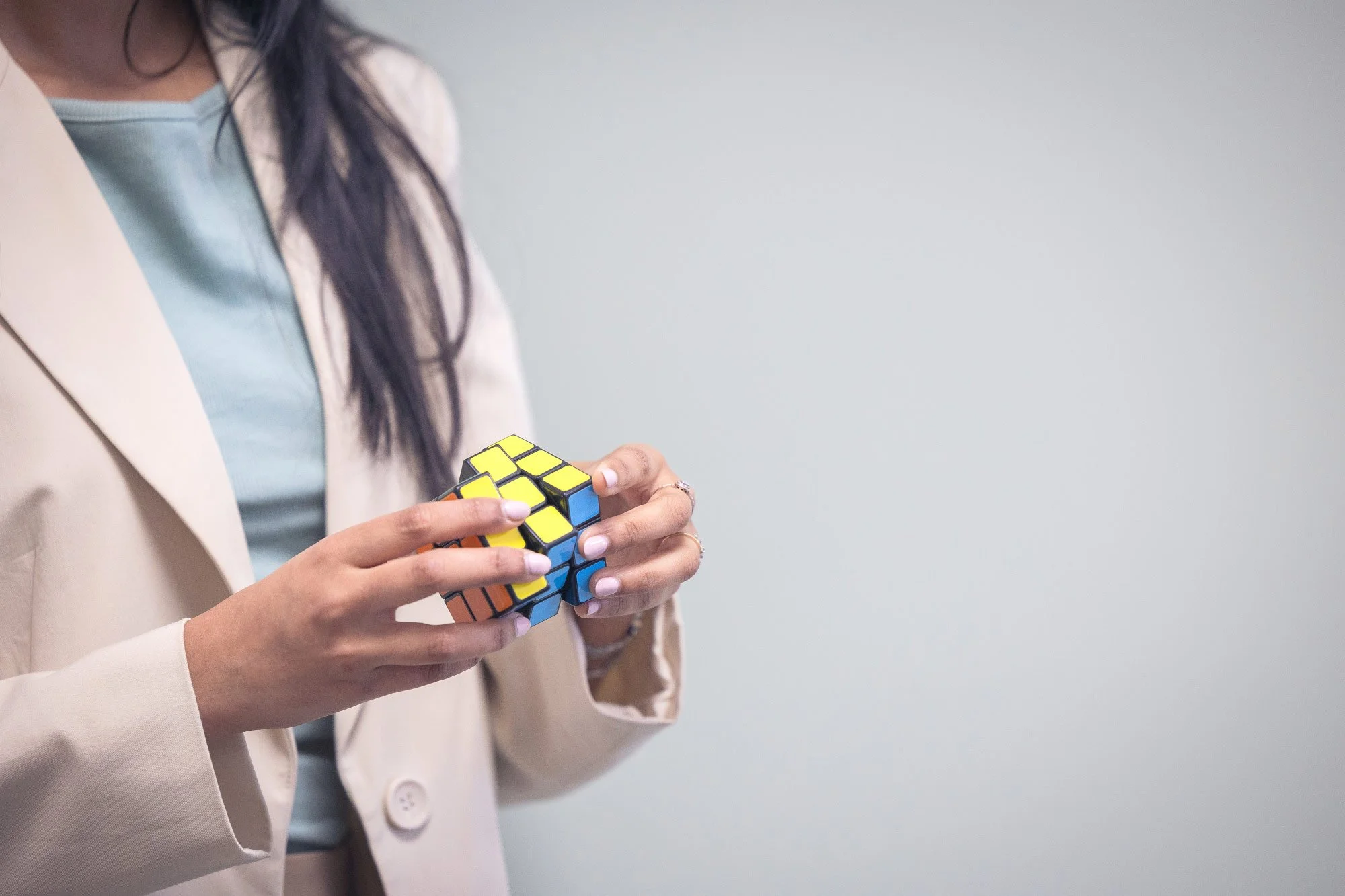 A woman wearing a face mask and beige blazer holding a colorful Rubik's Cube with both hands against a plain grey background.