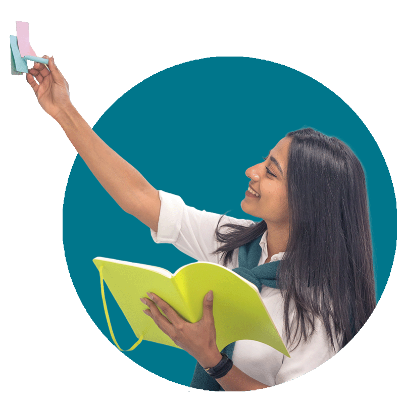 A young woman smiling and holding an open green book in one hand while reaching up to write on post it notes with her other hand.