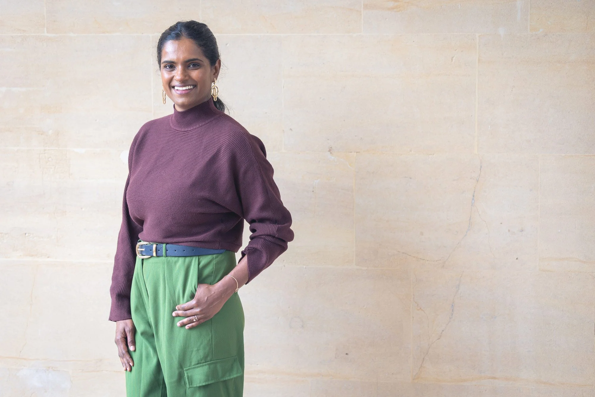 A woman with dark hair pulled back, wearing a maroon long-sleeved sweater with a high neckline, gold earrings, and a small bracelet, standing against a beige stone wall. She is smiling and has her right hand on her hip, wearing bright green pants with a blue belt.