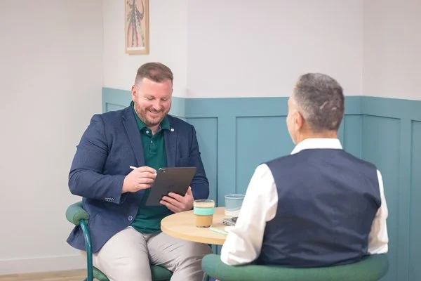 Two men sitting at a small round table in a room, engaged in conversation. One man is facing away from the camera, wearing a dark vest over a white shirt. The other man, facing the camera, is smiling and holding a tablet.