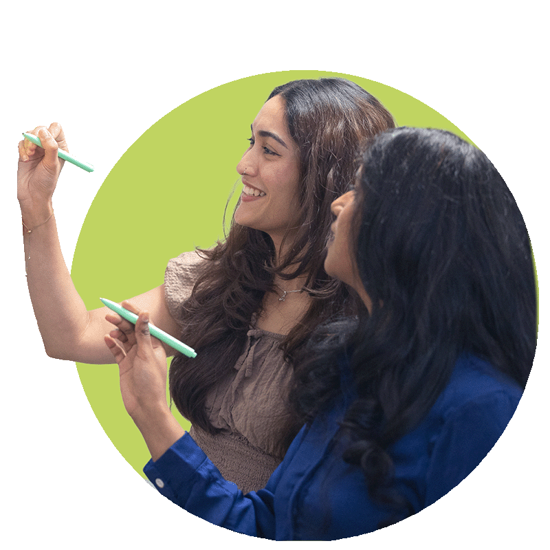 Two women with dark and light brown hair smiling and writing on a transparent surface with green pens, against a circular green background.