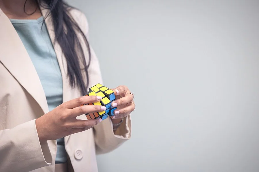 A woman in a beige blazer and light blue shirt holding a colorful Rubik's Cube against a plain gray background.
