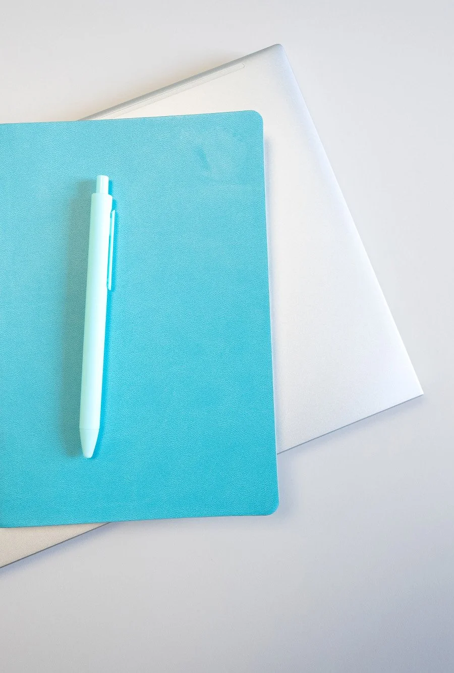 A pen placed on top of a blue notebook, with a silver laptop partially visible underneath on a white surface.