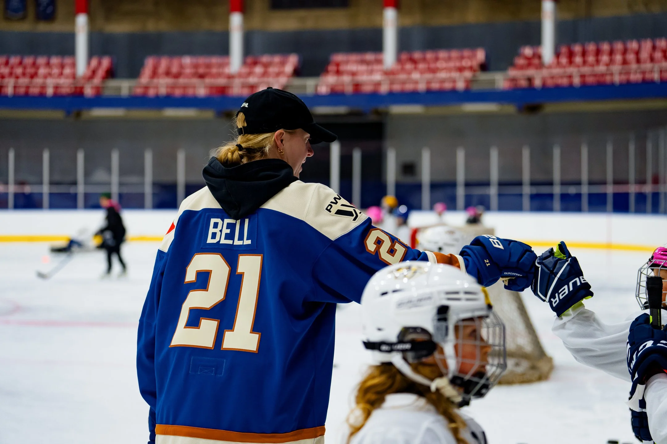 Hockey coach or player on ice rink wearing a jersey with the name BELL and number 21, guiding young players during practice or game, with other players and spectators in background.