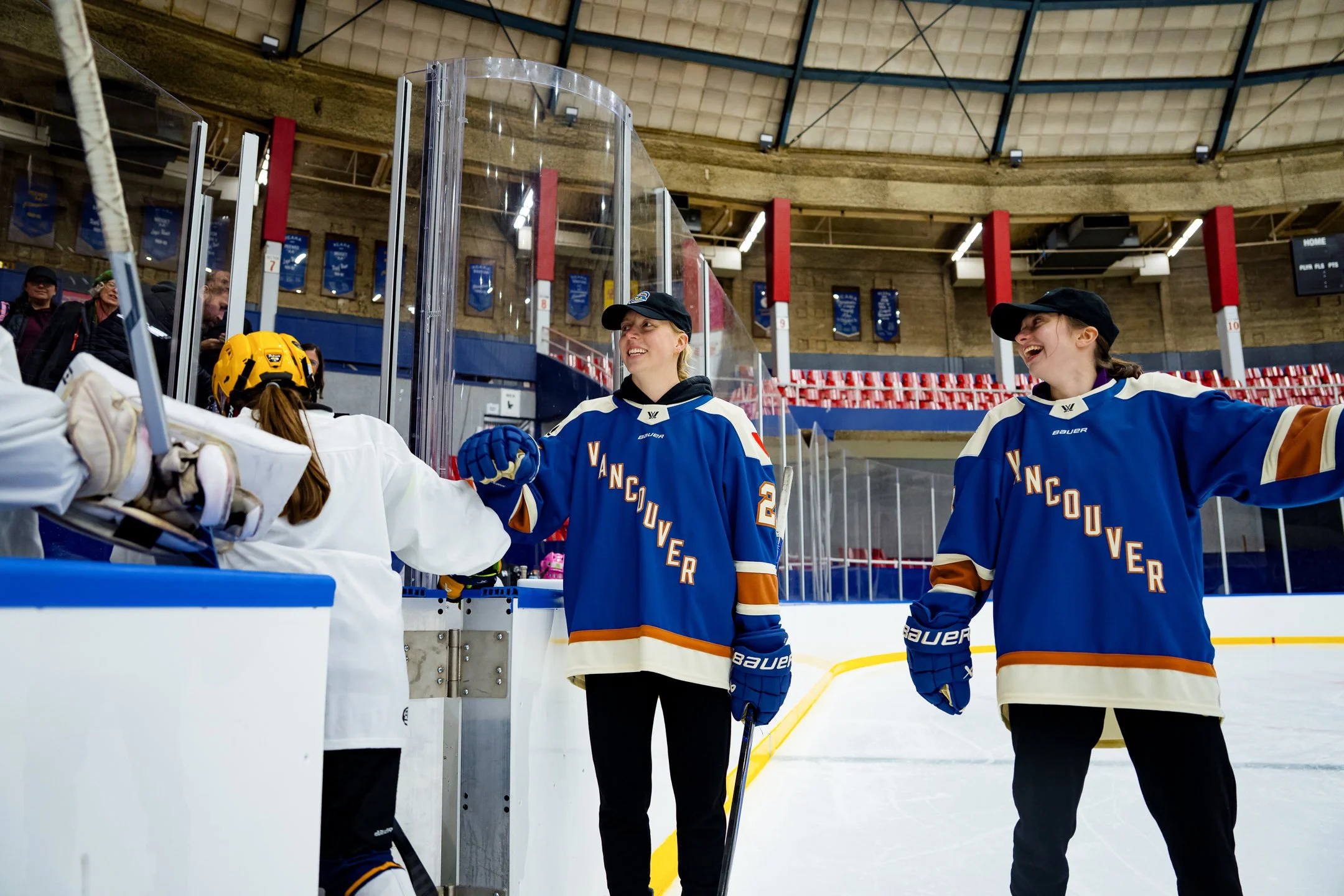 Two female hockey players in blue jerseys with orange and white accents, smiling and greeting each other on an ice rink. There are spectators in the background and a yellow helmet on the bench.