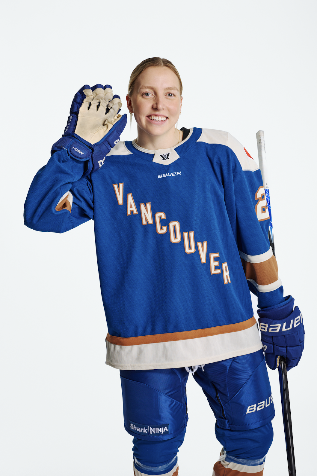 A female ice hockey player dressed in a blue Vancouver team jersey, with matching gloves and pants, holding a hockey stick, smiling and waving. She has blonde hair and a light complexion.
