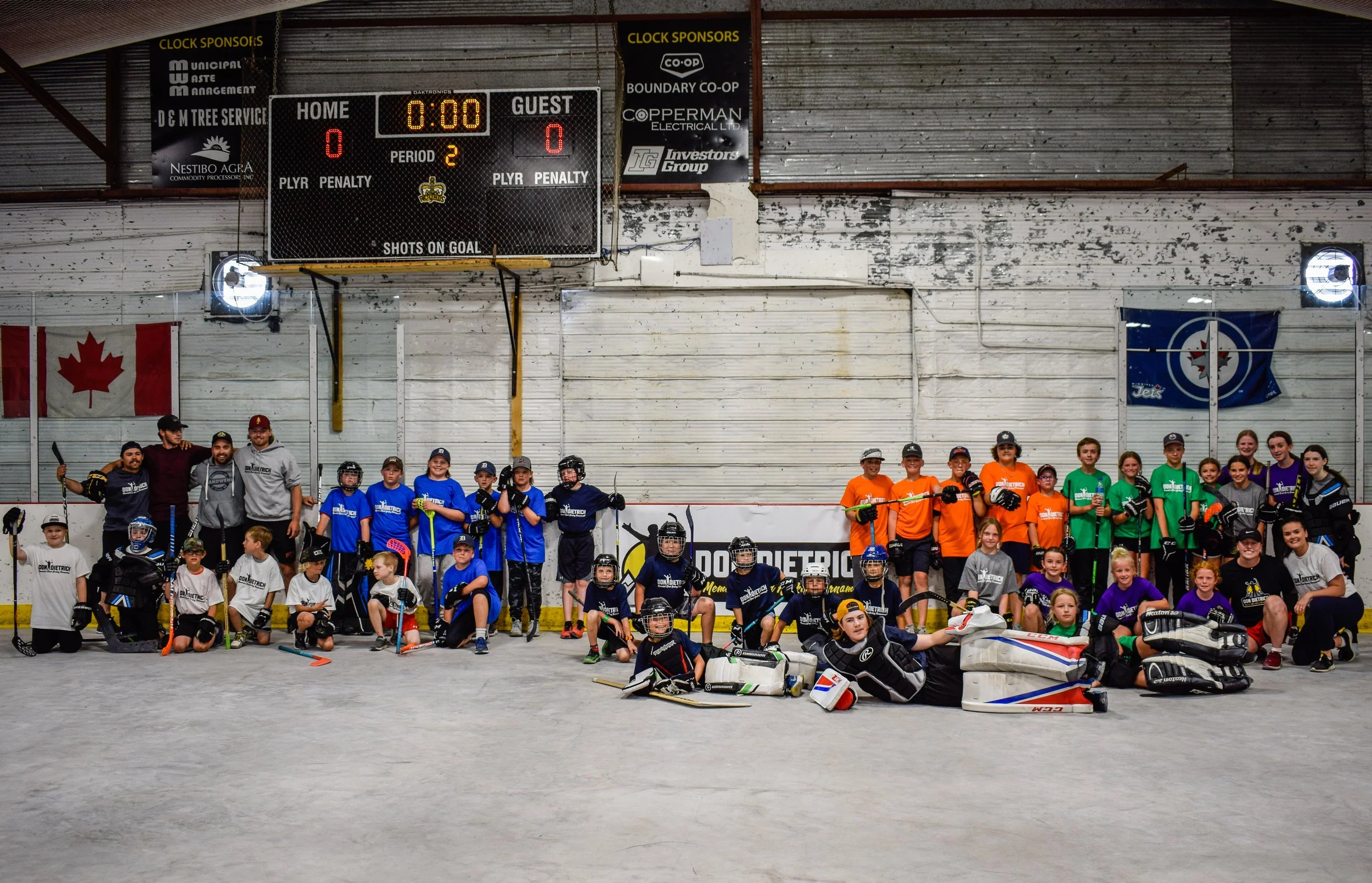 Group of children and coaches posing for a photo on an ice rink with hockey equipment, wearing various colored jerseys, in front of a digital scoreboard and hockey rink walls.