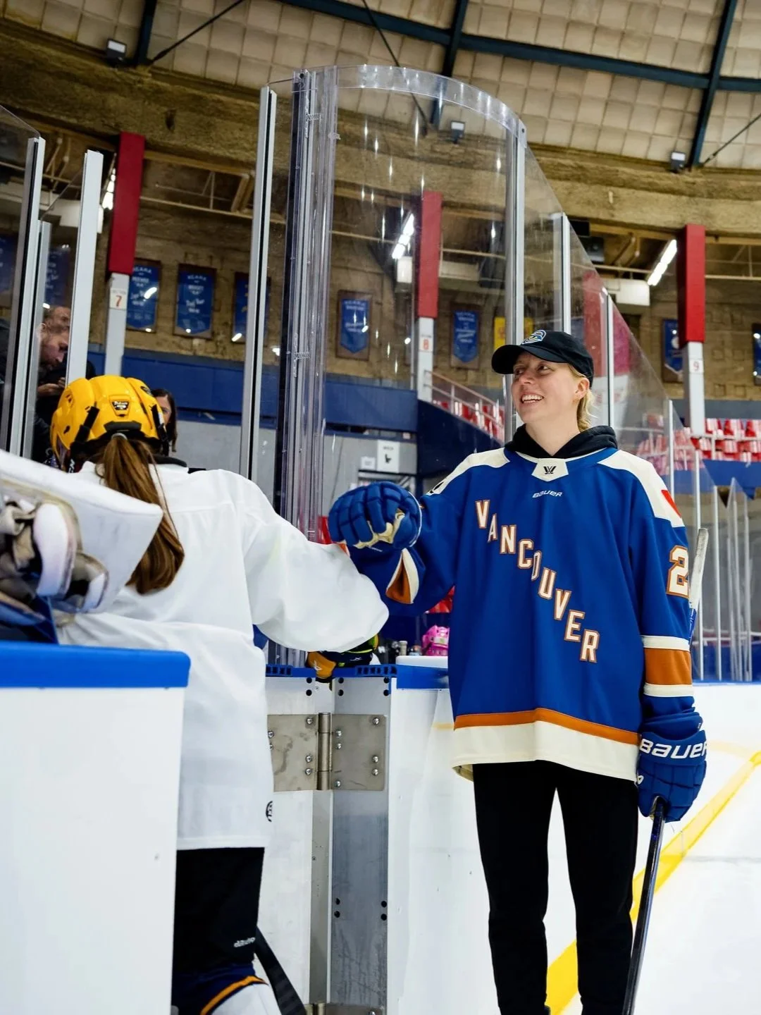 A female hockey player in a blue Vancouver jersey smiling and shaking hands with a teammate in a white jersey at an ice rink.