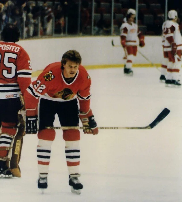 A hockey player in a red Chicago Blackhawks jersey, number 22, is bent over holding a hockey stick on the ice rink. Other players in red and white jerseys are visible in the background.