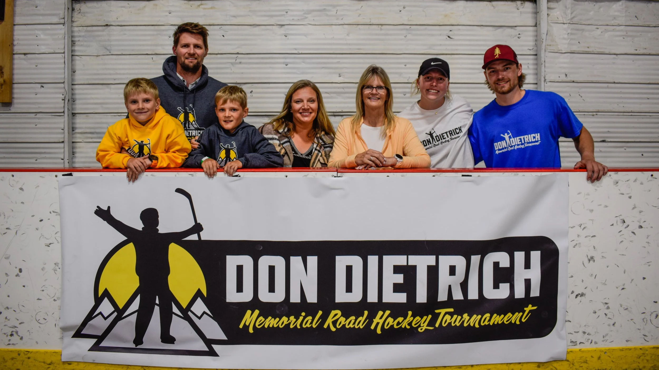 Group of seven people, including children and adults, posing behind a sign at a hockey tournament. They are inside an ice rink with a metal wall background. The sign reads 'DON DIETRICH Memorial Road Hockey Tournament' and features a silhouette of a child with a hockey stick.