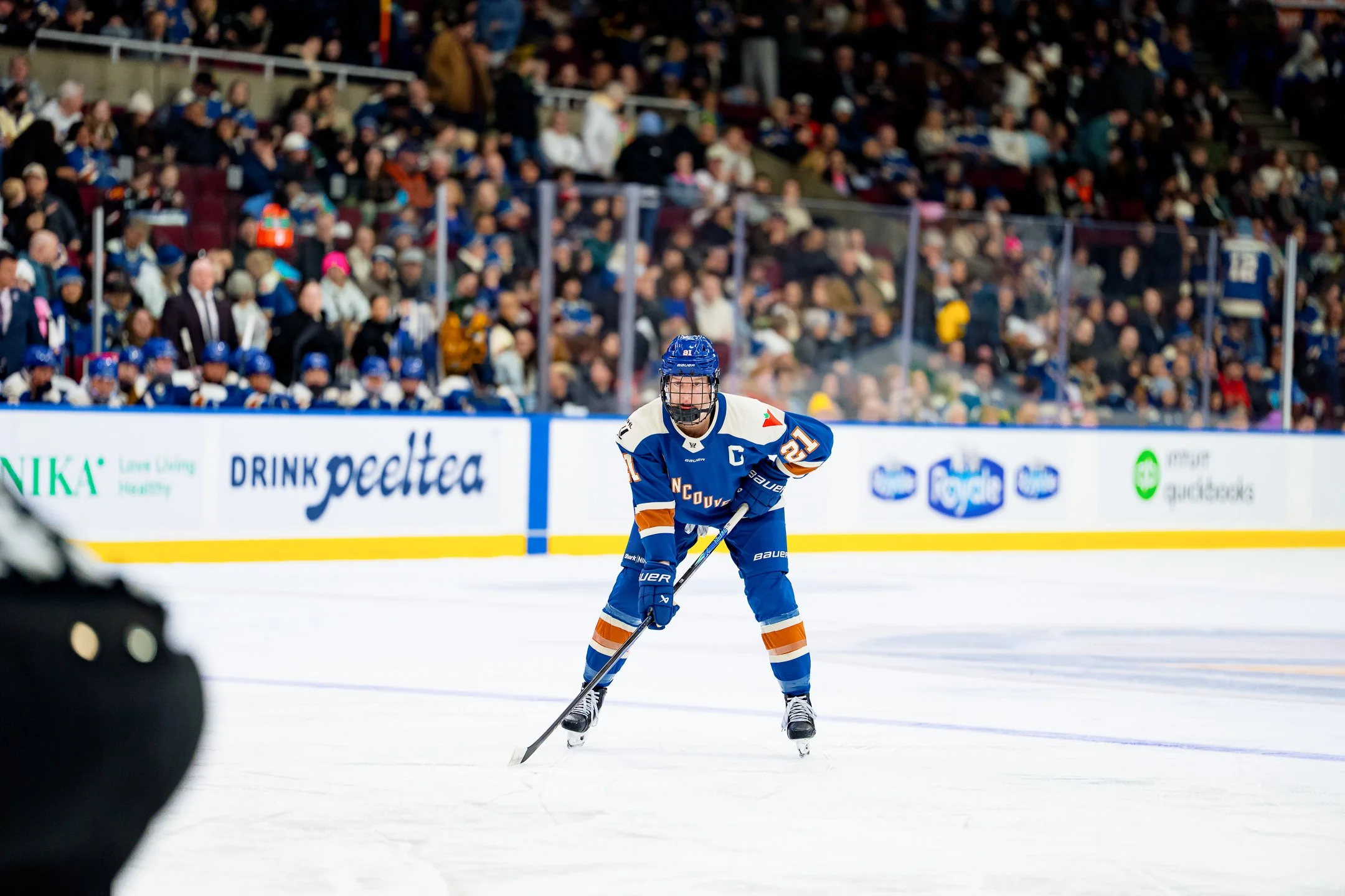 An ice hockey player in a blue and orange uniform, wearing a helmet and gloves, is skating on ice with a hockey stick during a game. A crowd of spectators is visible in the background in the stands.
