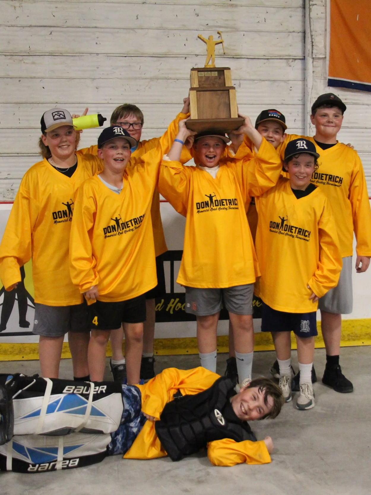 A group of young boys in yellow hockey jerseys celebrating after winning a tournament, holding a trophy, with one boy lying on the ground smiling at the camera.